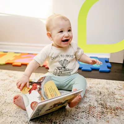 A smiling baby holding a book and wearing a beige onesie and sage green pants looks off camera.