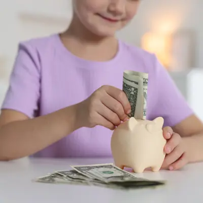 A young girl in a purple t-shirt putting money into a piggy banks.
