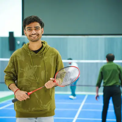 A young man in a green hoodie holds a badminton racket and stands on a blue badminton court.
