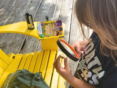 Child using a magnifying glass to inspect a rock standing beside a bright yellow Adirondack chair.