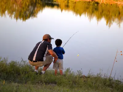 A man crouching beside a small child on a river bank. The child is holding a fishing rod.