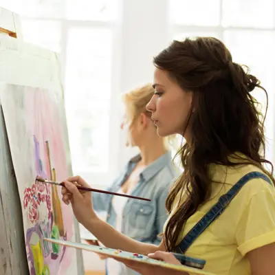 A woman in a yellow t-shirt painting on a canvas on an easel. 