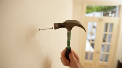A hand holding a hammer and hammering a nail into a cream coloured wall.