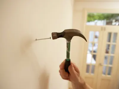 A hand holding a hammer and hammering a nail into a cream coloured wall.