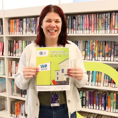 A woman holding the library's spring program guide standing in front of bookshelves.