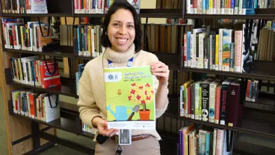 A female library staff member holds up a program booklet while smiling.