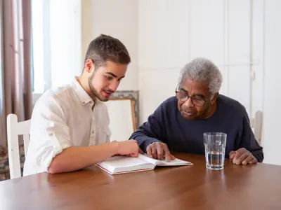A young man helping a senior man read a book while sitting at a table.