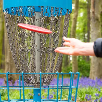 A hand tosses a red frisbee towards a blue metal disc golf basket.