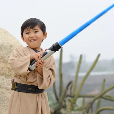 A young boy in a Jedi outfit holding a blue lightsaber toy.