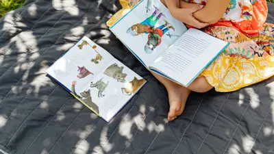 A child sitting on a black blanket on the grass reading with a book in her lap.
